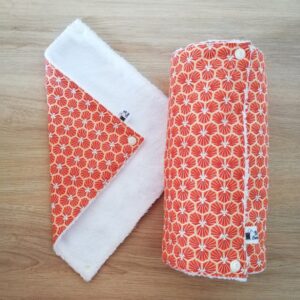 photo d'un rouleau d'essuie-tout, une face en éponge douce blanche et l'autre face en coton orange, motif géométrique. Les feuilles d'essuie-tout se fixent les unes aux autres à l'aide de boutons-pressions blancs