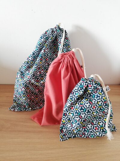 photo d'un lot de trois sacs à vrac (remplis) de tailles différentes, confectionnés dans un coton rose orangé pour la taille intermédiaire et coton géométrique multicolore pour le petit et le grand modèle. Les sacs à vrac se ferment à l'aide d'un cordon et d'une perle en bois