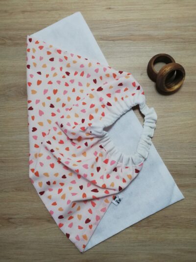 photo d'une serviette de table pour enfant, coton blanc uni d'un côté et rose orangé avec pour motif des cœurs de l'autre. Un élastique en haut de la serviette permet aux enfants de pouvoir la mettre facilement autour du cou.