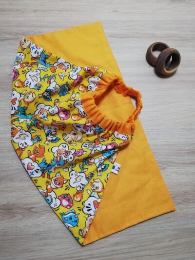 photo d'une serviette de table pour enfant, coton orange uni d'un côté et jaune avec pour motif des animaux inspiration manga. Un élastique en haut de la serviette permet aux enfants de pouvoir la mettre facilement autour du cou.